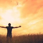 man with outstretched arms in field with sun